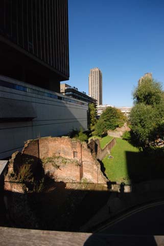 Ancient Roman wall is seen from a bridge crossing over from the Barbican to the Museum of London. The modern Museum building can be seen to the left with a tall sky scraper in the background.